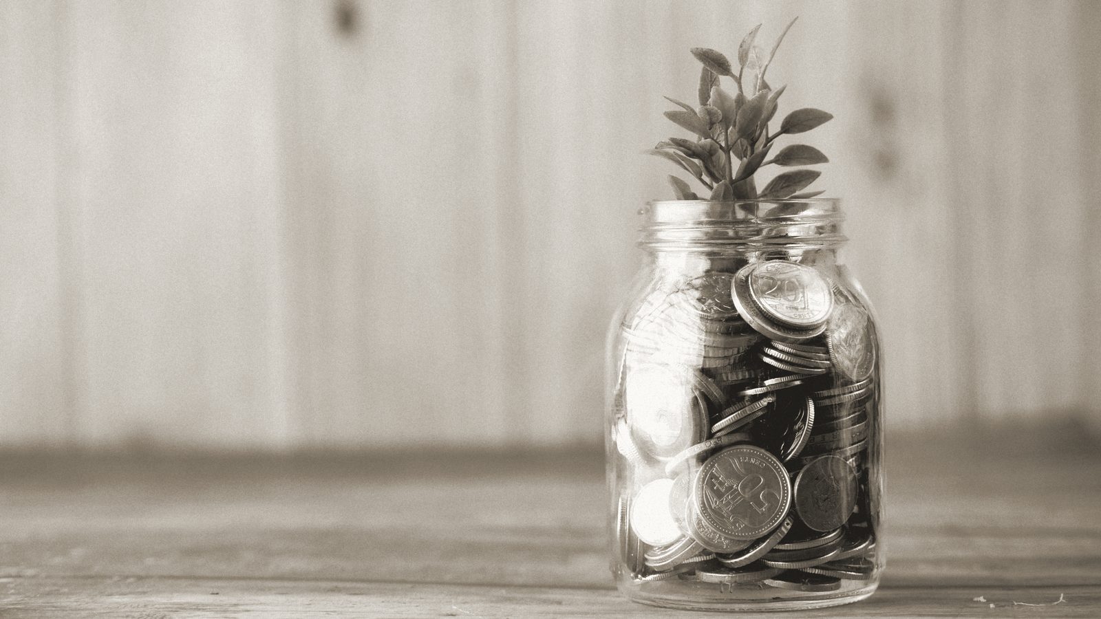 Un tarro de cristal lleno de monedas, del que crece una pequeña planta, sobre una mesa de madera